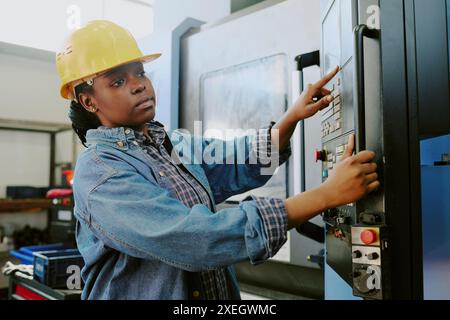 Medium shot of young African American woman wearing hardhat working in factory operating industrial CNC machine, copy space Stock Photo