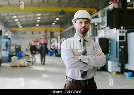 Medium portrait of confident Caucasian engineer wearing hardhat posing for camera with arms crossed at work in factory, copy space Stock Photo