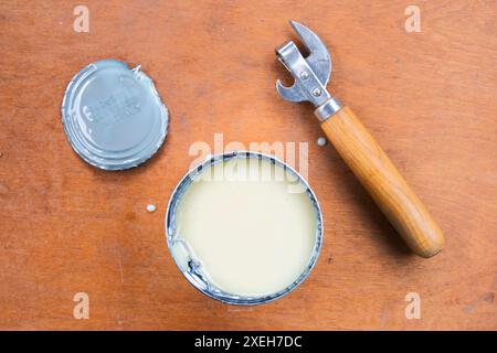 Open metal can with condensed milk, cut off lid, can opener on brown background, top view. Stock Photo