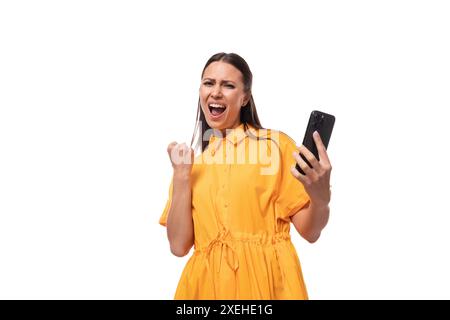 Young caucasian brunette woman with long hair dressed in a yellow summer dress rejoices in success holding a phone in her hand Stock Photo