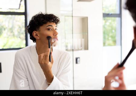 Applying makeup, transgender woman in bathrobe looking in mirror at home Stock Photo