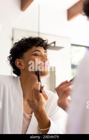 Applying makeup, transgender woman smiling in front of bathroom mirror at home Stock Photo