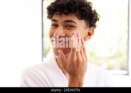 Applying face cream, young young man in bathrobe smiling in natural light Stock Photo