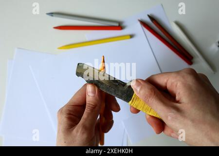 Photo of process of sharpening colored pencils with a clerical knife. Human hands. Yellow pencil. Metal knife. On the background are white papers and Stock Photo