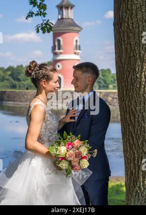 Young beautiful bride and groom stand in front of the water against the backdrop of a pink lighthouse. Newlyweds on a walk. Wedding, love, married cou Stock Photo