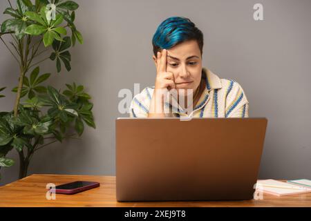 Concentrated androgynous person with blue hair working on laptop - thoughtful and focused, LGBT pride bracelet, modern home office setup, inclusive an Stock Photo