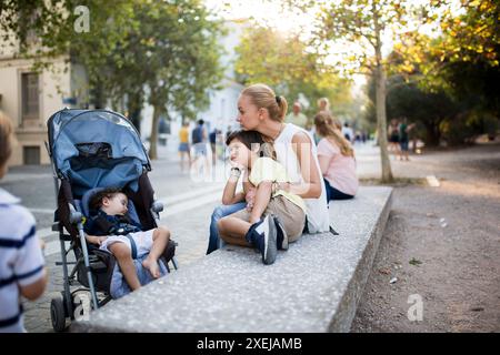 A mother and her two sons enjoy a leisurely moment outdoors in the city, with one son sleeping in a stroller and the other leaning against his mother. Stock Photo