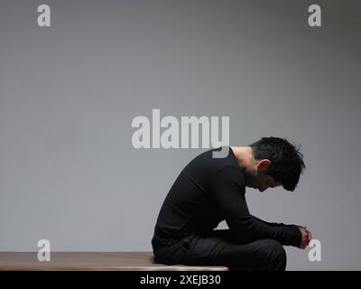 A Hispanic man dressed in black sits with hands clasped and head down. Stock Photo