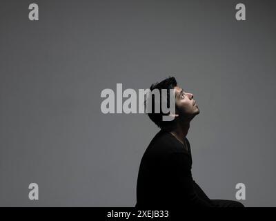 A Hispanic man dressed in black sits in front of a gray background and looks up. Stock Photo