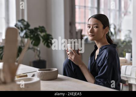 Asian female artist sitting at craft table in ceramic studio relaxing Stock Photo