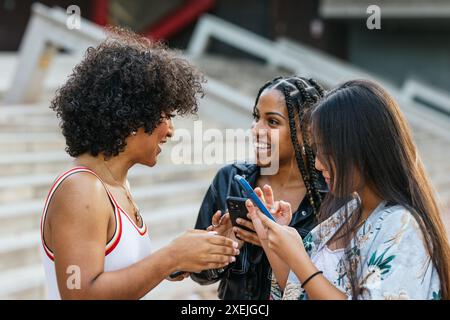 Three young women sharing laughs and browsing phone outdoors on city steps. Stock Photo