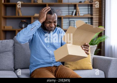 Man sitting on couch in living room looking at open empty box with confused expression. Concept of disappointment, confusion, online shopping issue, unexpected delivery, or missing item. Stock Photo