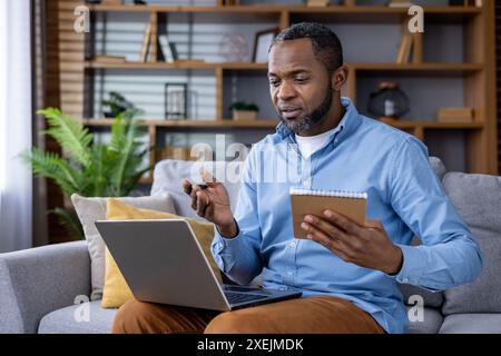 Man sitting on couch at home working remotely using laptop and holding notebook. Engaged in video call, appearing focused and thoughtful, surrounded by modern home office decor. Stock Photo