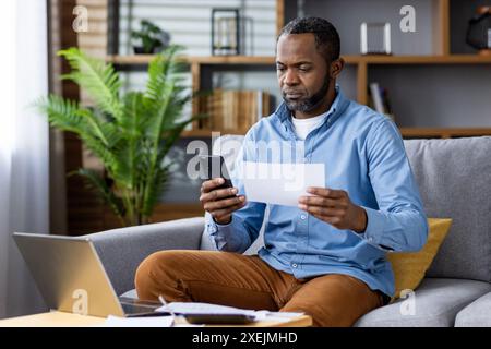 Man working from home, multitasking with smartphone, laptop, and documents, focusing on remote work in a modern living room setting with plants and bookshelves Stock Photo
