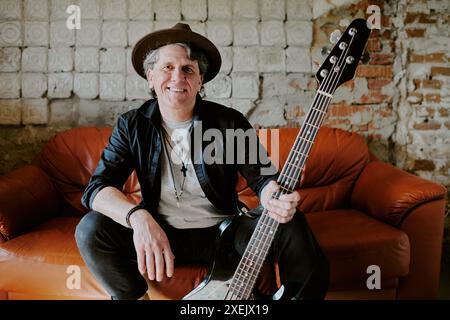 Medium portrait of confident mature man wearing hat sitting on leather sofa holding bass guitar and posing for camera Stock Photo