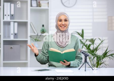 Smiling woman in hijab giving online lecture holding book, speaking into microphone. Bright modern office background. Concept of education, communication, public speaking, teaching online Stock Photo