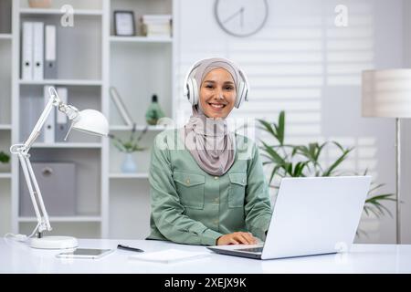 Smiling woman wearing hijab working on laptop at home office with headphones. Bright and modern workspace featuring white furniture, bookshelf, and plants. Concept of remote work and productivity Stock Photo