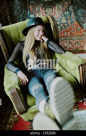 Vertical high angle view portrait of gen Y female grunge musician wearing hat sitting in old green armchair and smiling at camera Stock Photo