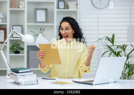 Happy woman celebrating after receiving good news in letter while working from home office. Expression of joy and success with laptop and books on the desk. Stock Photo