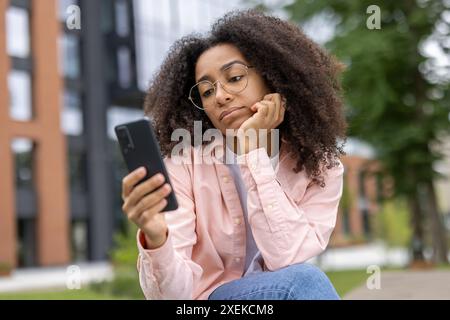 Young female student looking bored while using smartphone near university campus building. Concept of technology, boredom, and student life. Stock Photo