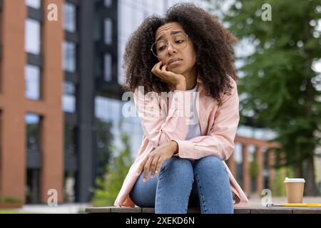 Young woman with curly hair and glasses sitting on bench in urban park, looking bored or sad. Casual attire with a coffee cup and notebook nearby. Stock Photo
