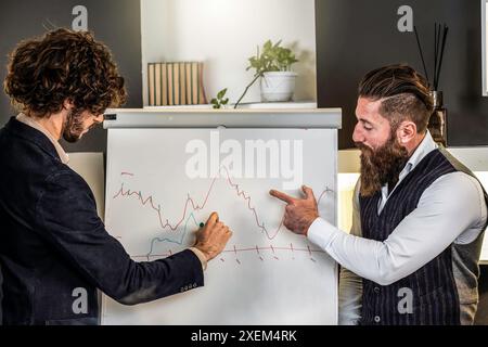 Businessmen analyzing financial chart on whiteboard. Two colleagues discussing data trends during a business meeting. Modern office environment. Stock Photo