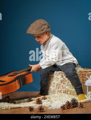 A little boy plays the guitar on a blue background. Nearby are pine cones and a candlestick. The child is wearing a light shirt and a cap on his head. Stock Photo