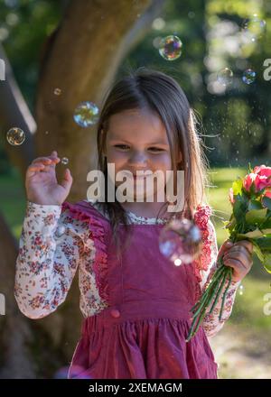 Close-up portrait of a little cute girl. A child holds a small bouquet of roses. Soap bubbles fly nearby. Joy, delight, happy childhood Stock Photo