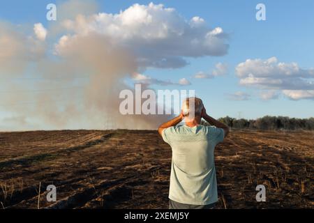 A man, who has his back turned, medium shot, looks at his burned field. He has his hands above his head. There is still smoke from the fire at the bot Stock Photo