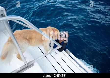 Service dog interacts with a paraplegic man in the ocean water at the back of a boat, ready to scuba dive in a wetsuit and goggles Stock Photo