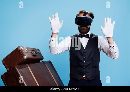 Modern doorman using vr glasses in studio, wearing formal suit and tie to work at luxurious five star hotel. Bellhop with 3d virtual reality headset presenting his professionalism. Stock Photo