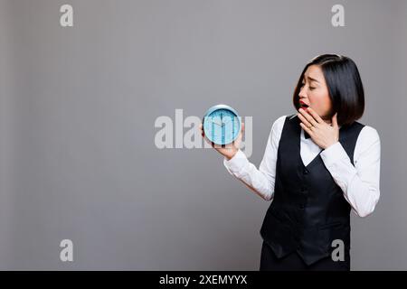 Asian waitress overslept and running late for work at cafe while yawning and looking at alarm clock. Restaurant sleepy woman employee wearing professional uniform showing time Stock Photo