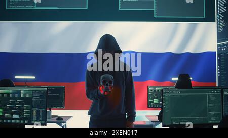 Conceptual image of a hooded russian hacker creating hazardous alerts, holds a skull and warning of death threats. Russian federation works on cybercrime and violence, panic and torment. Camera B. Stock Photo