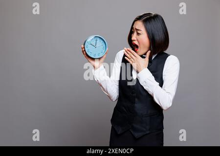 Sleepy asian waitress yawning and covering mouth with hand while looking at alarm clock. Tired young woman receptionist wearing restaurant uniform checking time while working late Stock Photo