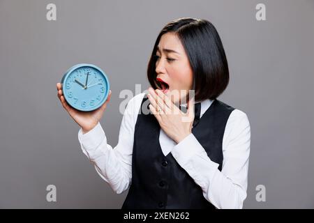 Tired overslept asian waitress in uniform holding circle dial alarm clock and checking time. Restaurant sleepy woman receptionist looking at round retro watch while yawning Stock Photo