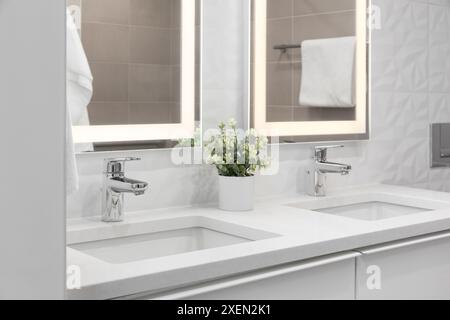 A bathroom detail with chrome faucets on a white marble countertop and cabinet, pattern tiled wall, and vanity mirrors with lights. Stock Photo
