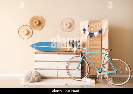 Surfboard on modern chest of drawers and bicycle near beige wall in room Stock Photo