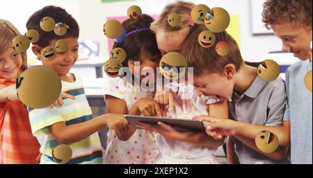 Image of emoticons floating over diverse children laughing and using digital tablet in classroom Stock Photo