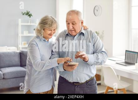 Senior couple standing in living room at home using mobile phone for texting or social media. Stock Photo