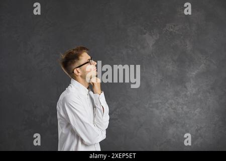 Pensive young man standing on gray background and looking at copy space touching his chin. Stock Photo