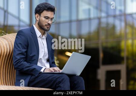 Businessman working on laptop outdoors in professional attire. Modern office building in background. Professional, focused, and engaged in work. Represents productivity, modern work environment Stock Photo