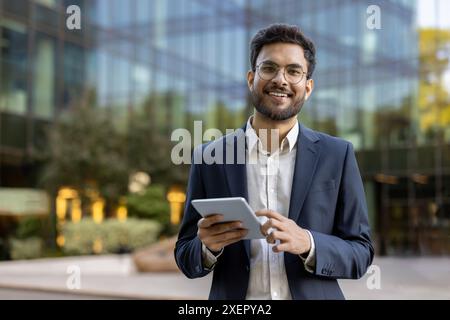 Young businessman in suit using tablet outside modern glass office building. Confident professional smiling working on digital device. Concept of business, technology, success, and entrepreneurship. Stock Photo