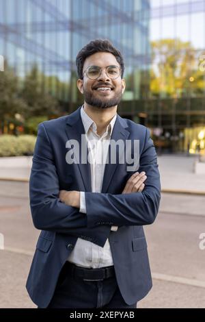 Confident businessman standing with arms crossed outside modern glass office building, expressing success and leadership in corporate environment Stock Photo