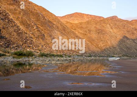 The barren mountains forming the edge of the Fish River Canyon, gold in the early morning light, reflecting into the river and pools Stock Photo