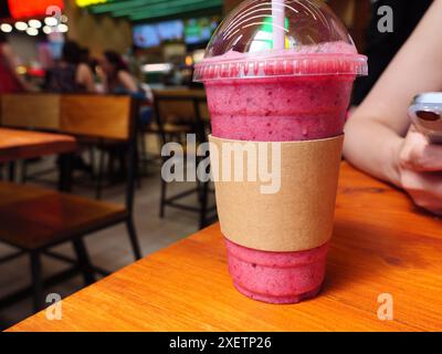 A pink smoothie in a plastic cup sits on a wooden table in a Cafe. The smoothie cup has an empty paper cup sleeve on it. Carton cup sleeve mockup. Stock Photo