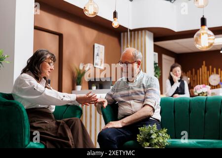 Caucasian retired old husband and wife sharing their vacation ideas in luxurious lounge area. Elderly couple sitting and chatting in hotel reception waiting for accommodation, ready for fun honeymoon. Stock Photo