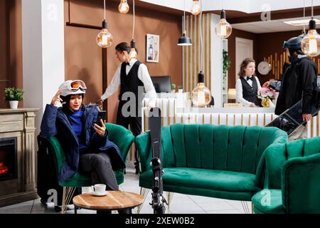 Joyful asian lady having video call on her cell phone in lounge area of snowy mountain resort. Youthful female guest, surrounded by ski gear, participates in video conversation using mobile device. Stock Photo