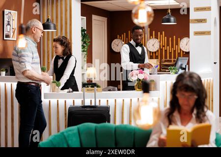 Senior male tourist arrives at front desk of hotel reception, where receptionists assists him with check-in process. Multiethnic employees help guest while elderly woman relaxes in luxury lounge area. Stock Photo