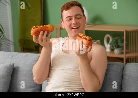 Young happy overweight man with tasty hot dogs sitting on sofa at home. Weight gain concept Stock Photo