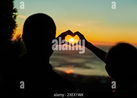 Sunset sky, silhouette and couple with heart hand, emoji or gesture on romantic tropical beach adventure together. Sun, island and date night for man Stock Photo